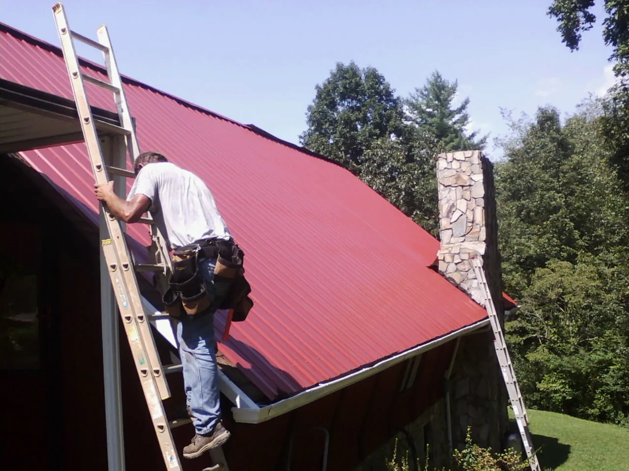 Roofer on a ladder beside a red metal roof and stone chimney — Soffit & Fascia Repair by Roofing Brothers in New Braunfels
